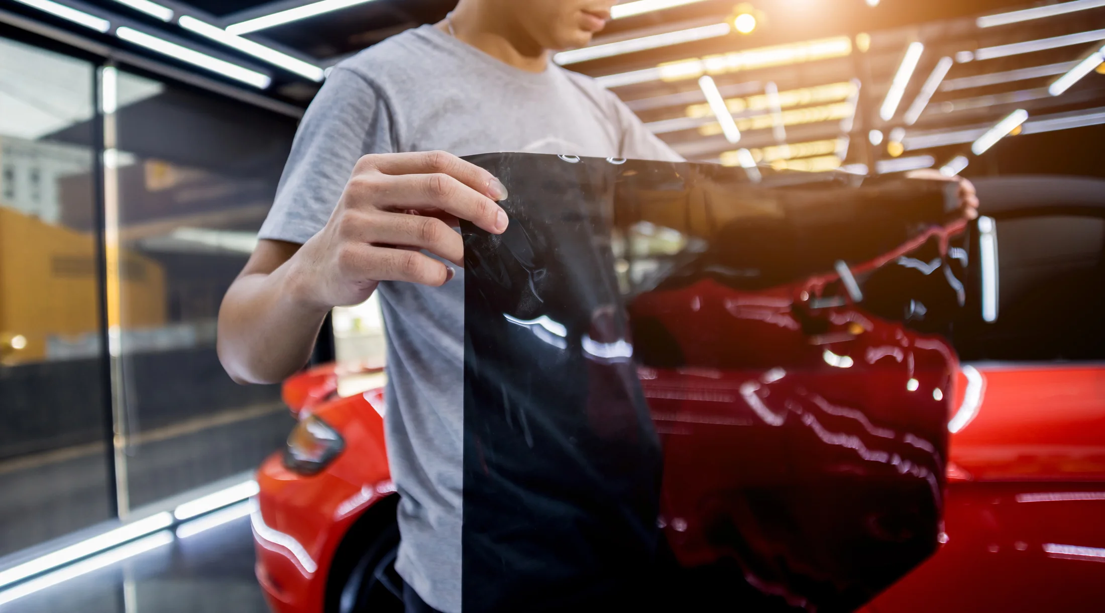 A tint installer preparing window film beside a red car in the workshop.