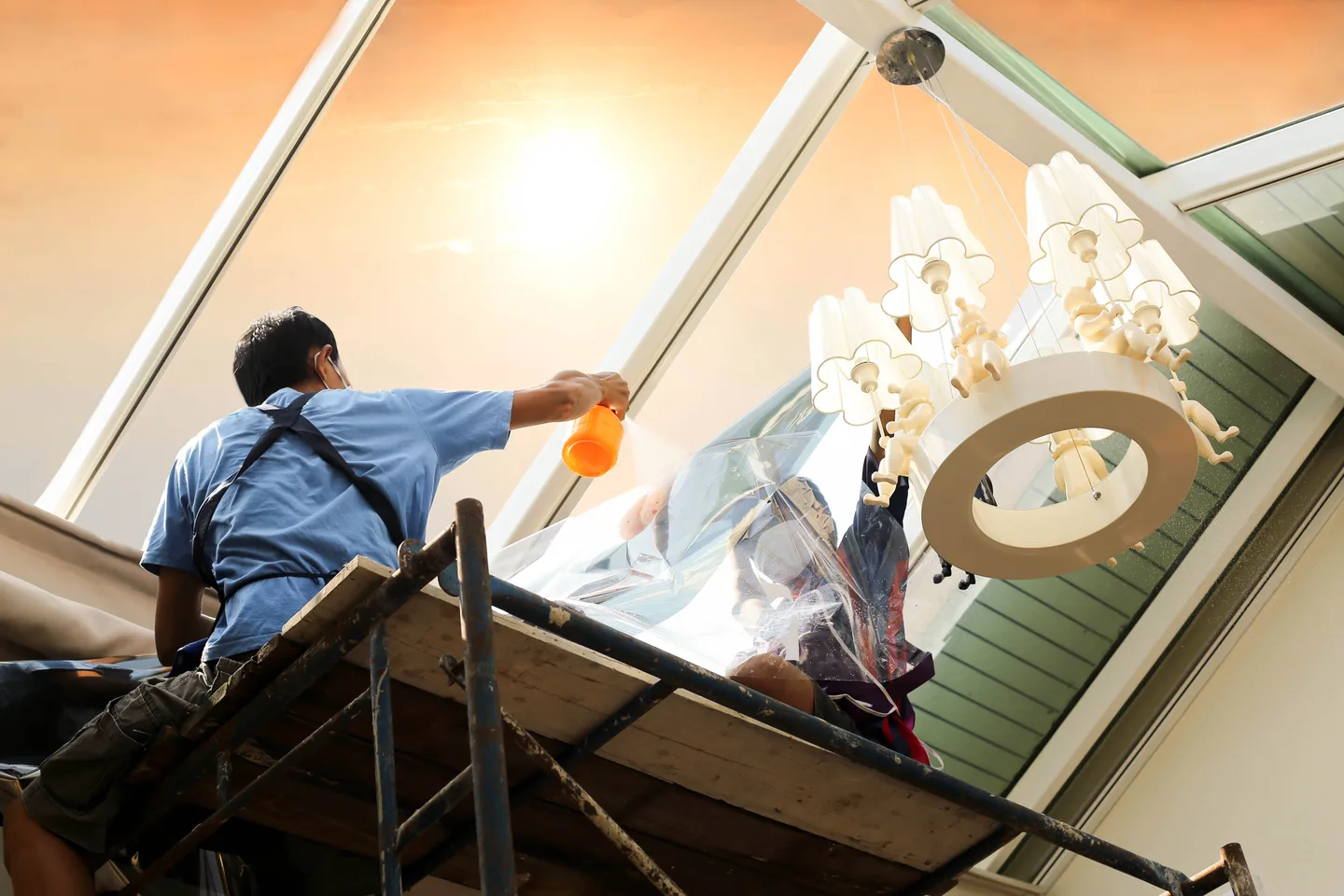 Installers applying window film beneath a sunlit glass roof, with a chandelier beside the work area.